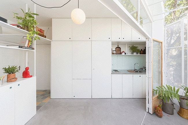 Ảnh chụp bên trong ngôi nhà nhỏ, cận cảnh khu bếp là một hệ tủ màu trắng có khoảng trống nhỏ đặt bếp và bồn rửa, bên trái là kệ để đồ nội thất nhà nhỏ