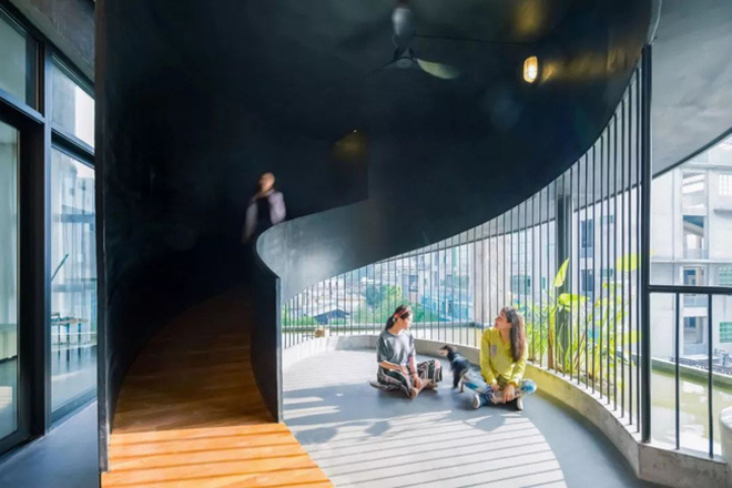 Cầu thang màu tối dẫn xuống lối đi bên phải là một sảnh ban công đầy nắng, nơi có hai người phụ nữ và một chú cún nhỏ đang ngồi thư giãn ban công nhà đẹp